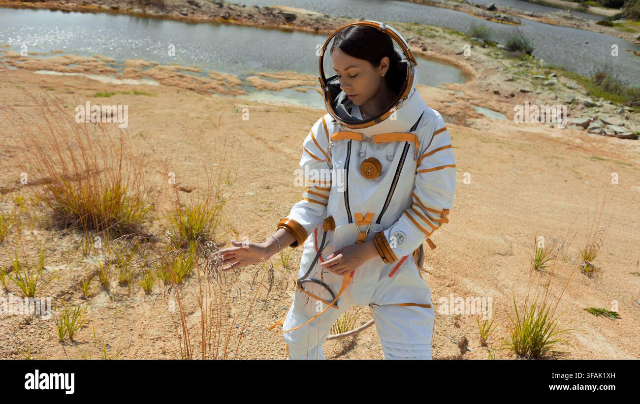 Exploratrice féminine se penche vers une végétation clairsemée sur un sol couleur rouille, illustrant l'écologie et l'équilibre fragile d'un monde habitable nouvellement découvert Banque D'Images