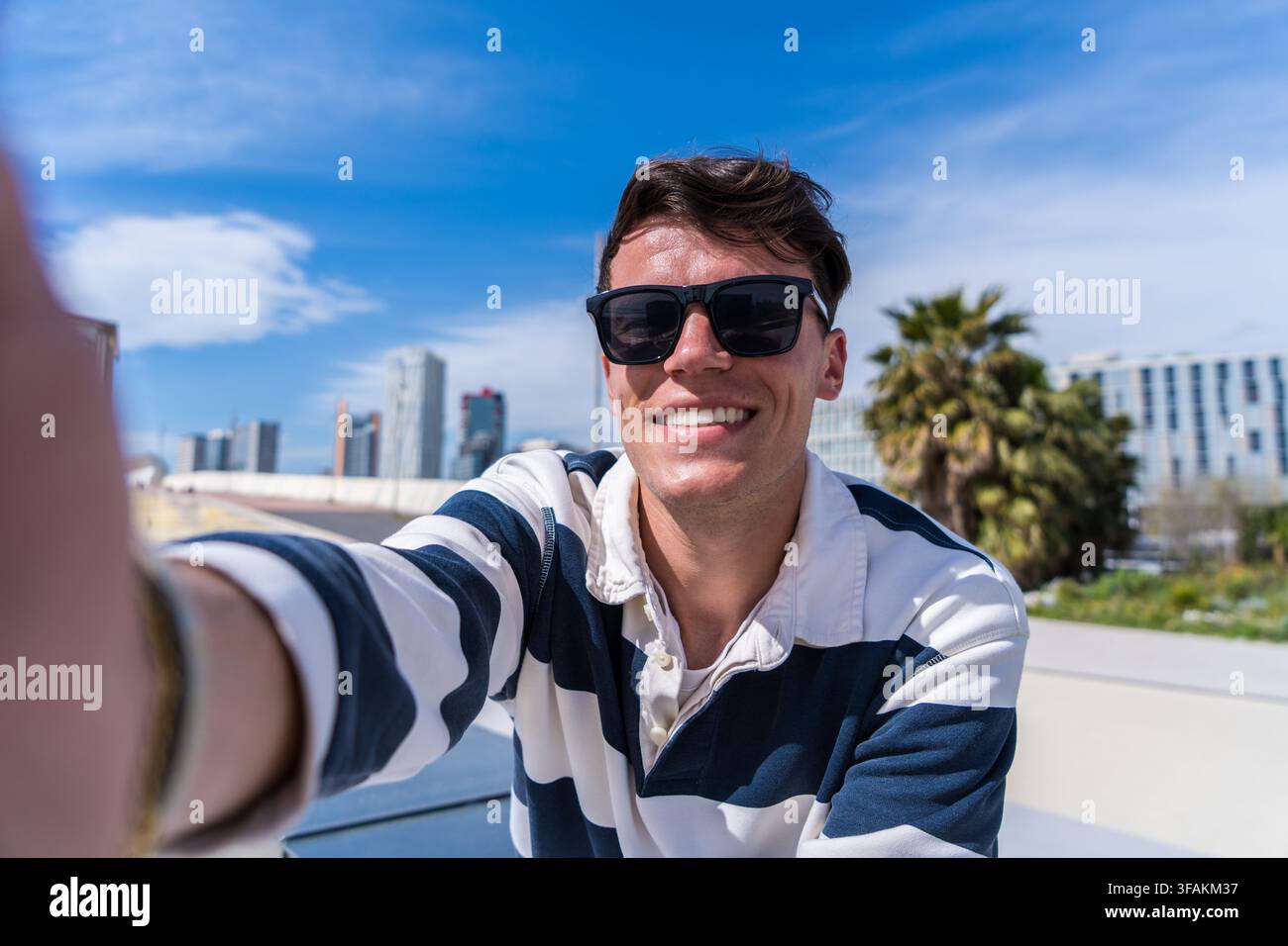 Jeune homme portant des lunettes de soleil prenant un selfie souriant en plein air dans une ville moderne avec un ciel bleu et des palmiers Banque D'Images