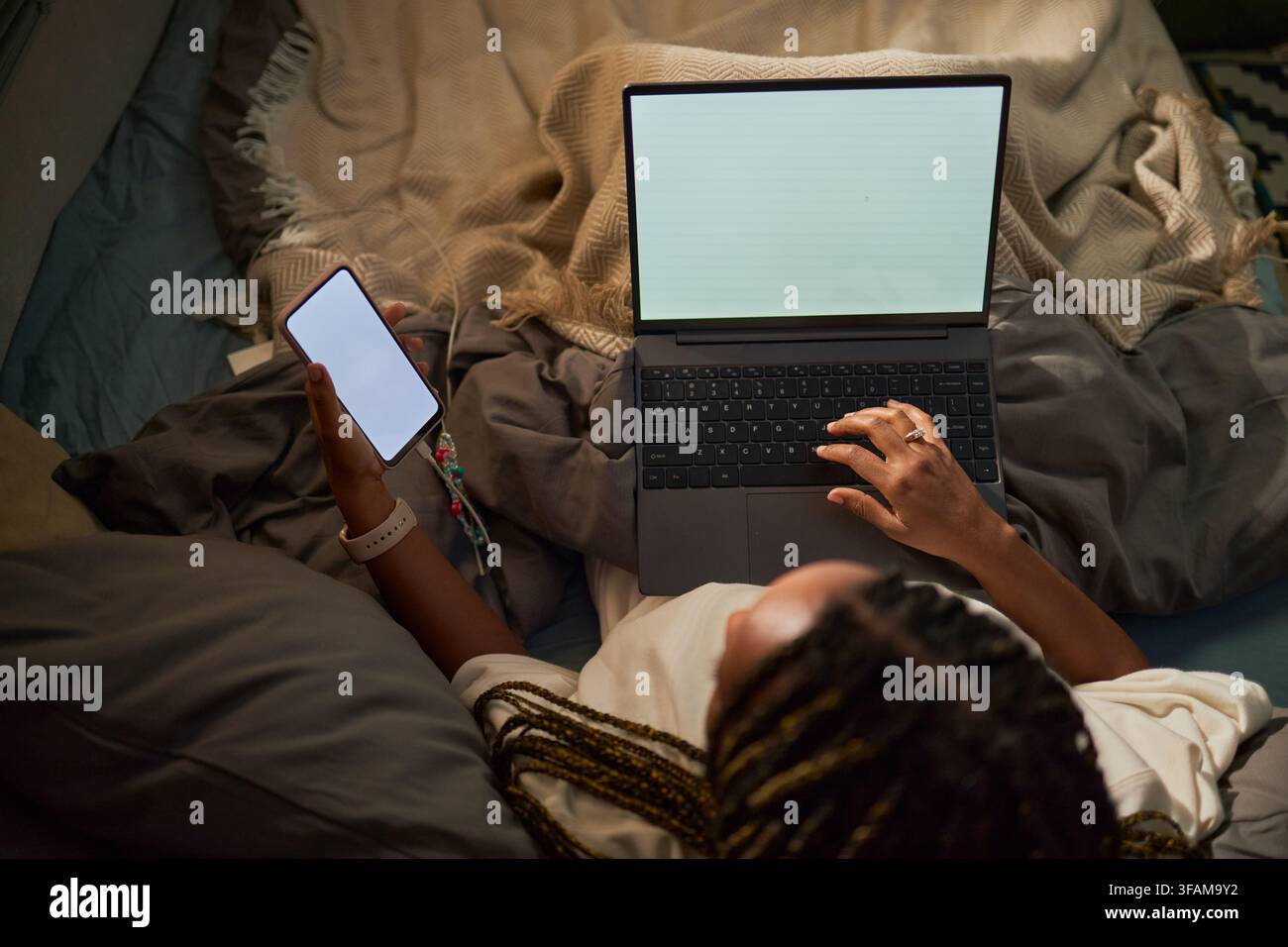 Femme noire multitâchant avec un ordinateur portable et un smartphone au lit Banque D'Images
