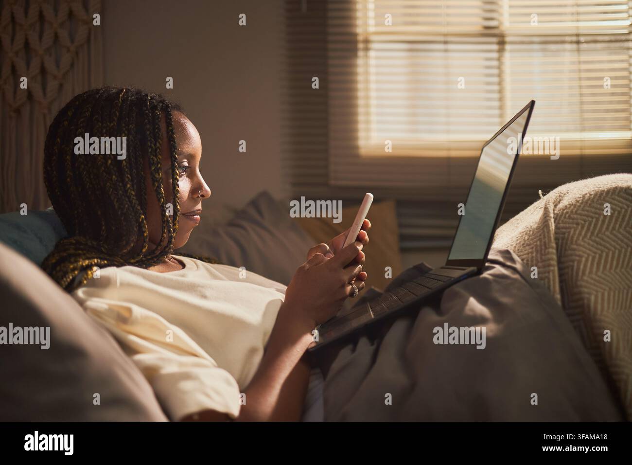 Femme noire utilisant un smartphone et un ordinateur portable au lit pour le travail à distance Banque D'Images