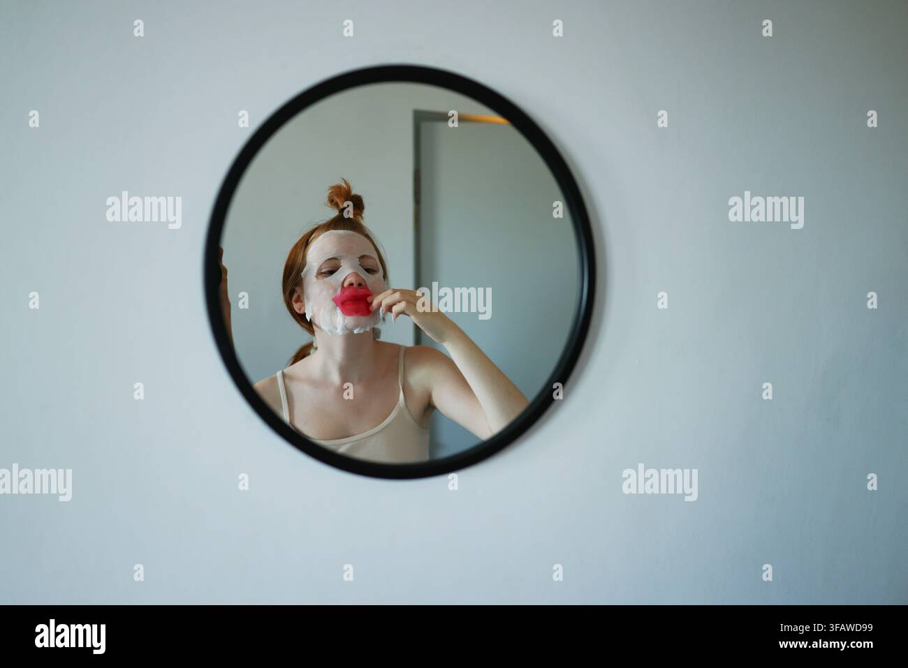 Jeune femme appliquant un masque en feuille et un patch pour les lèvres tout en se regardant dans le miroir pendant sa routine de soins à domicile. Banque D'Images