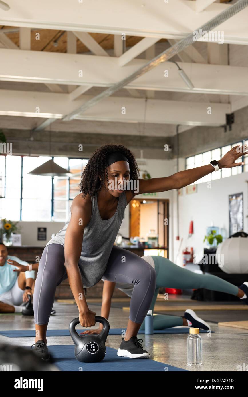 Classe de fitness afro-américaine effectuant un squat avec kettlebell de 5 kg, portant un haut gris sur un tapis dans le studio. Salle de sport, poids, groupe, mode de vie, industriel, motivation Banque D'Images