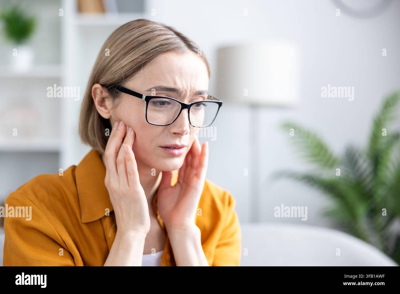 Femme mûre avec des lunettes touchant sa joue et grimçant à cause d'une douleur sévère aux dents ou à la mâchoire, stressée et mal à l'aise à l'intérieur d'une clinique dentaire ou lors d'un rendez-vous médical, gros plan Banque D'Images