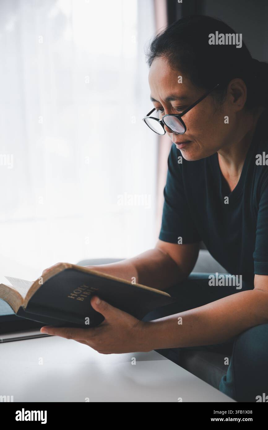 Femme d'âge moyen pensive portant des lunettes et lisant la Sainte Bible près d'une fenêtre, représentant la dévotion chrétienne, l'étude spirituelle, la foi, la sages Banque D'Images