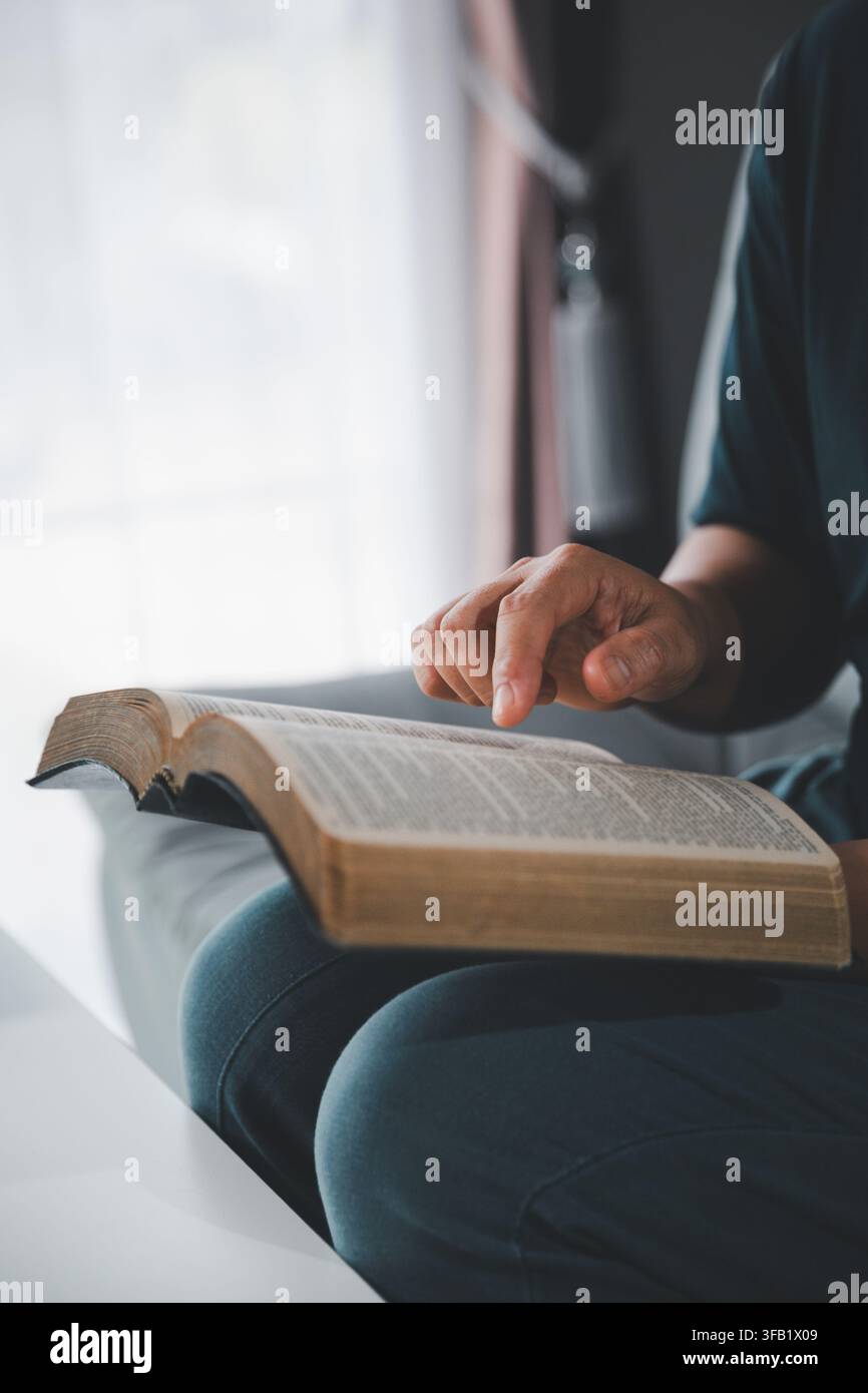 Gros plan latéral d'une femme suivant le texte avec son doigt tout en lisant une Bible ouverte près d'une fenêtre, symbolisant l'étude des Écritures, la foi, la sagesse, Banque D'Images