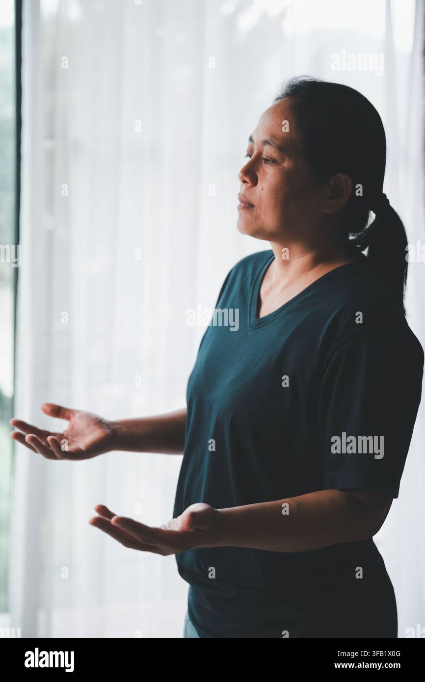 Vue latérale d'une femme d'âge moyen debout à côté d'une fenêtre lumineuse les mains ouvertes pendant la prière, exprimant l'adoration, la reddition, la gratitude, l'espoir et les tr Banque D'Images