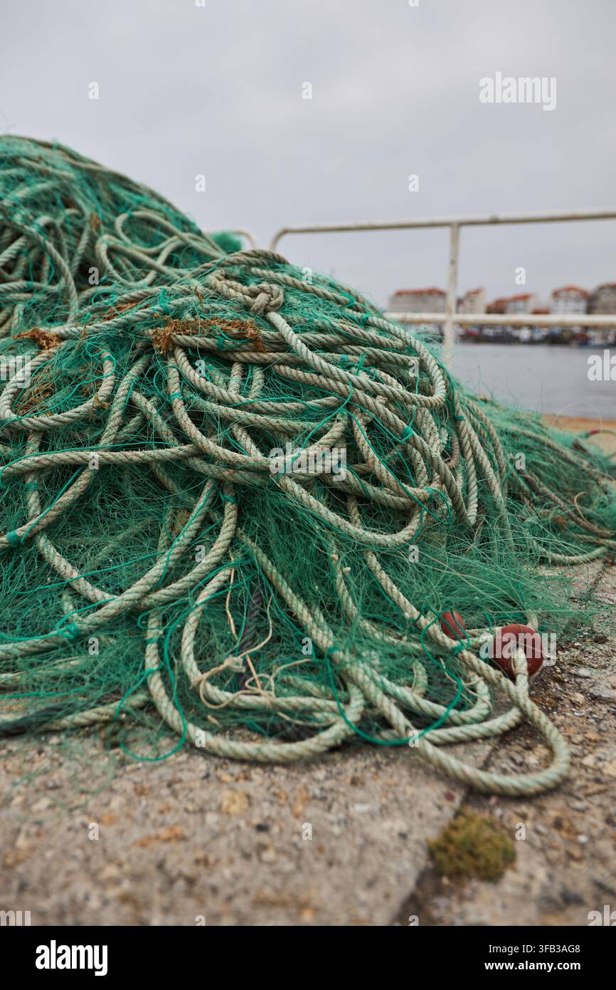 Close-up d'un filet de pêche vert emmêlé et d'une épaisse corde empilés sur un quai de port usé, avec de l'eau calme, la rambarde d'un bateau et des navires au loin sous un o Banque D'Images