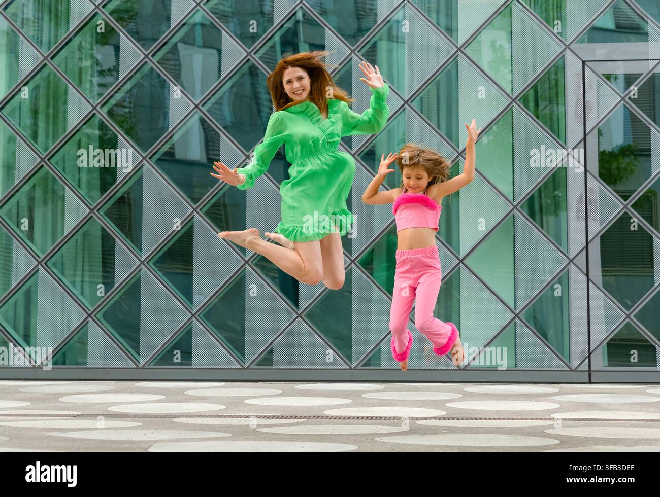 Mère et fille joyeuses sautent en l'air devant un bâtiment moderne en verre, portant des tenues vert vif et rose lors d'une journée estivale joyeuse dans le Banque D'Images