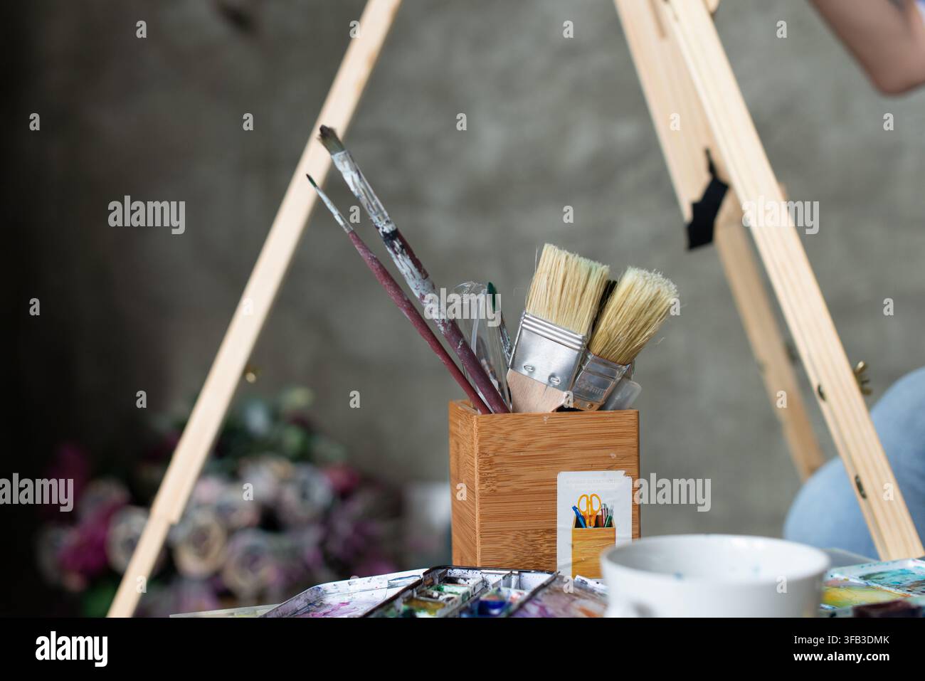 Gros plan de pinceaux à aquarelle, de couteaux à palette et de fournitures de peinture dans un porte-outils en bois à côté d'un chevalet, capturant l'atmosphère calme d'un artiste Banque D'Images