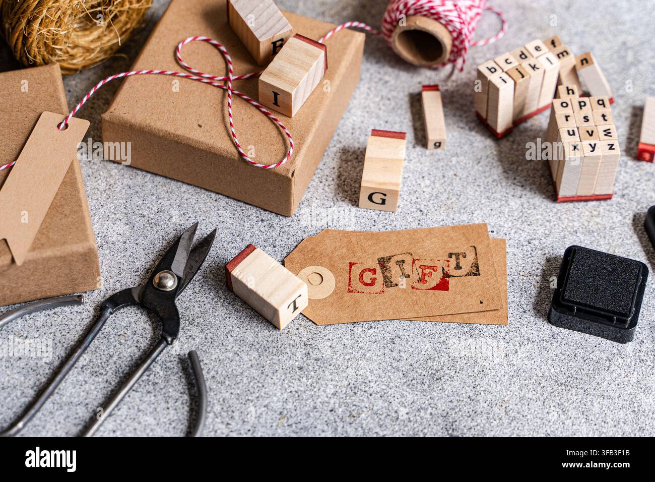 Vue aérienne de matériaux d'emballage cadeaux faits à la main avec des boîtes en papier kraft, des tampons alphabet, du fil rouge, des étiquettes vierges, un tampon encreur et des ciseaux sur une texture grise Banque D'Images