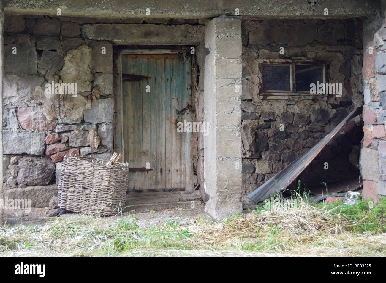 Une porte en bois patinée, une petite fenêtre et un panier en osier se dressent contre le mur de pierre croulant d'une ferme rurale abandonnée avec de l'herbe à son entr Banque D'Images