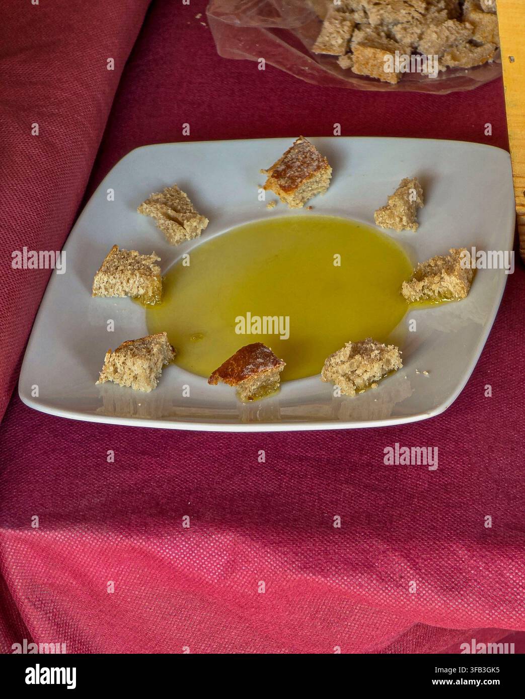 De petits morceaux de pain croustillant sont disposés autour d'une mare d'huile d'olive sur une assiette blanche carrée. Cette mise en place est destinée à une dégustation lors d'un événement gastronomique local, invitant Banque D'Images