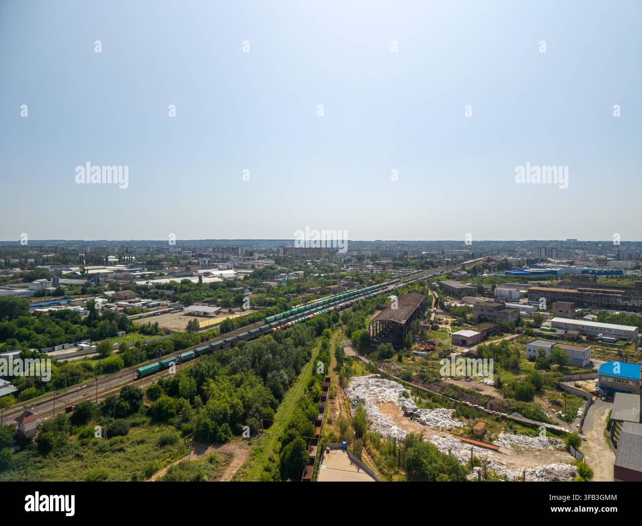 Une prise de drone en grand angle capture un long train de marchandises sur les voies ferrées entouré d'arbres verts, de bâtiments industriels, de structures anciennes et d'un vaste ciel urbain Banque D'Images