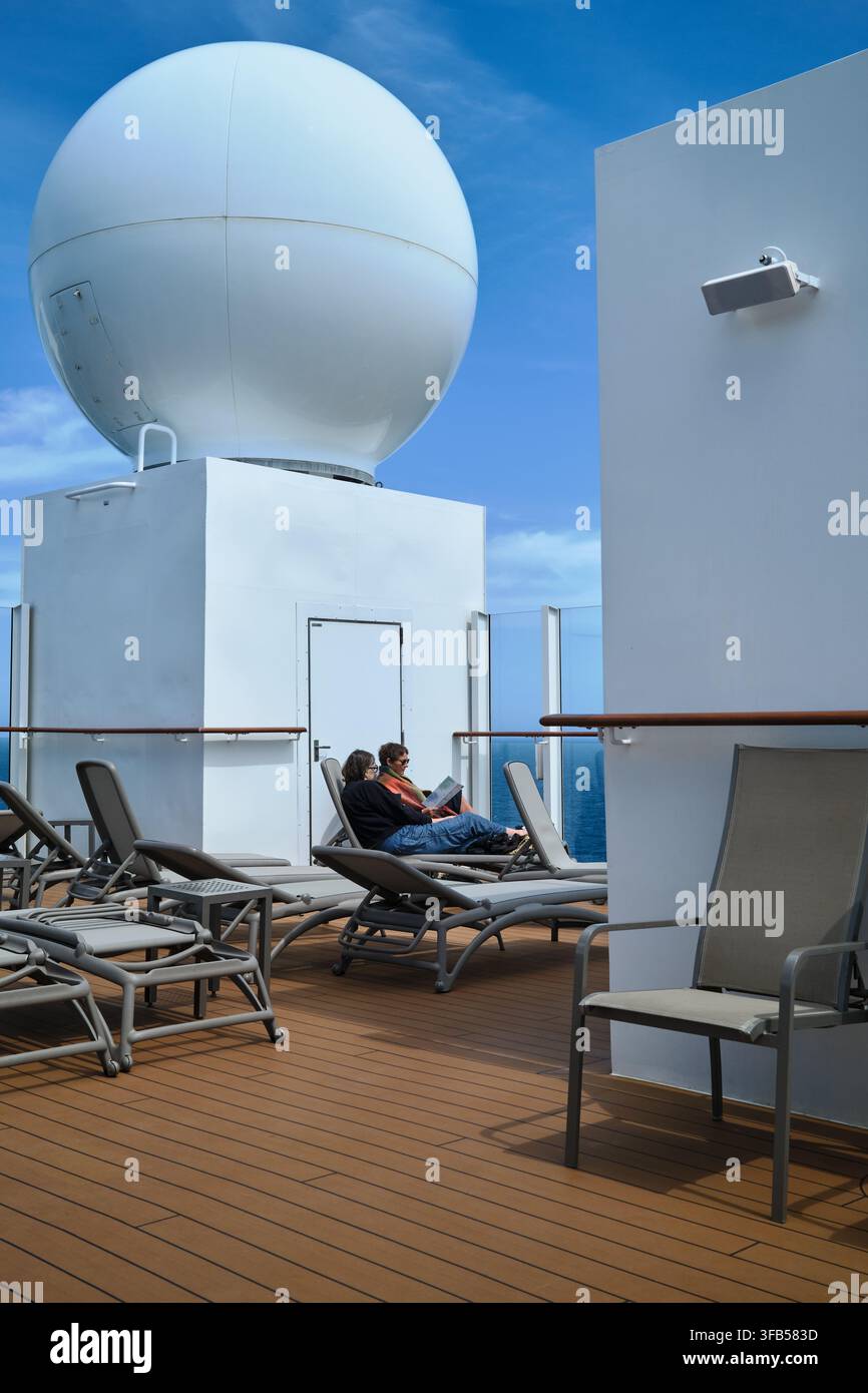 Passagers se détendant sur des chaises longues à côté d'un grand dôme blanc de communications par satellite sur le pont supérieur du navire de croisière MS Iona. Banque D'Images
