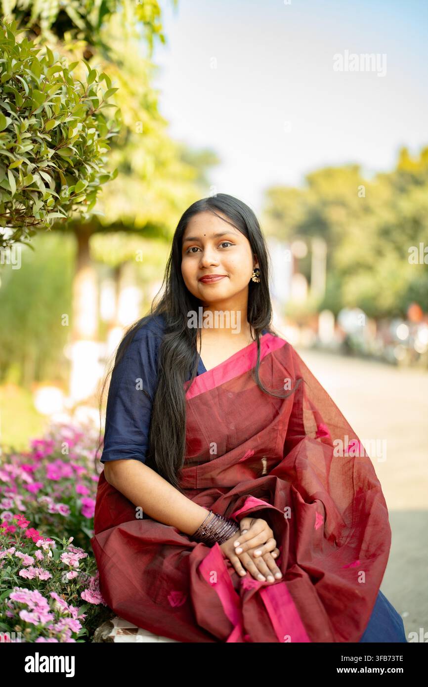 Une femme portant un saree marron avec un chemisier bleu est assise à l'extérieur près de plantes en fleurs et de verdure pendant la journée. Banque D'Images