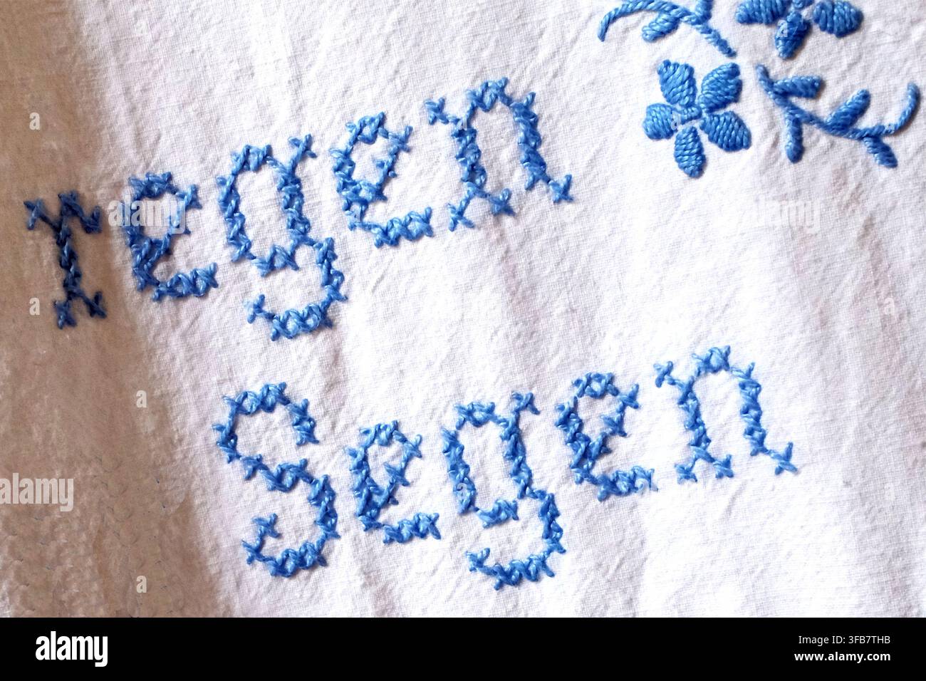 Tissu blanc avec broderie au point de croix bleu présentant les mots « regen » (pluie) et « Segen » (bénédiction) Le point de croix est dans une écriture décorative, compl Banque D'Images