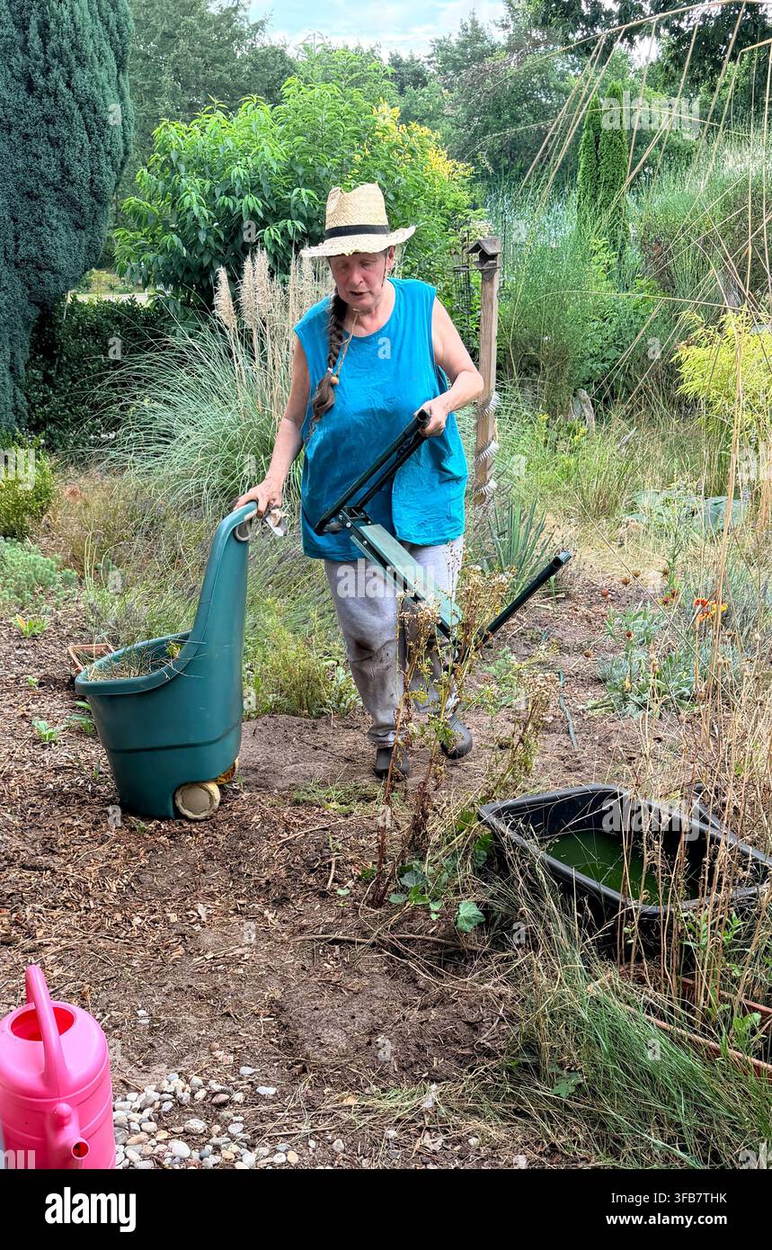 Une femme âgée travaille dans un jardin, portant un chapeau de paille, un haut bleu sans manches et un pantalon gris. Elle tient un genouillère de jardin et pousse une roue bleu-vert Banque D'Images