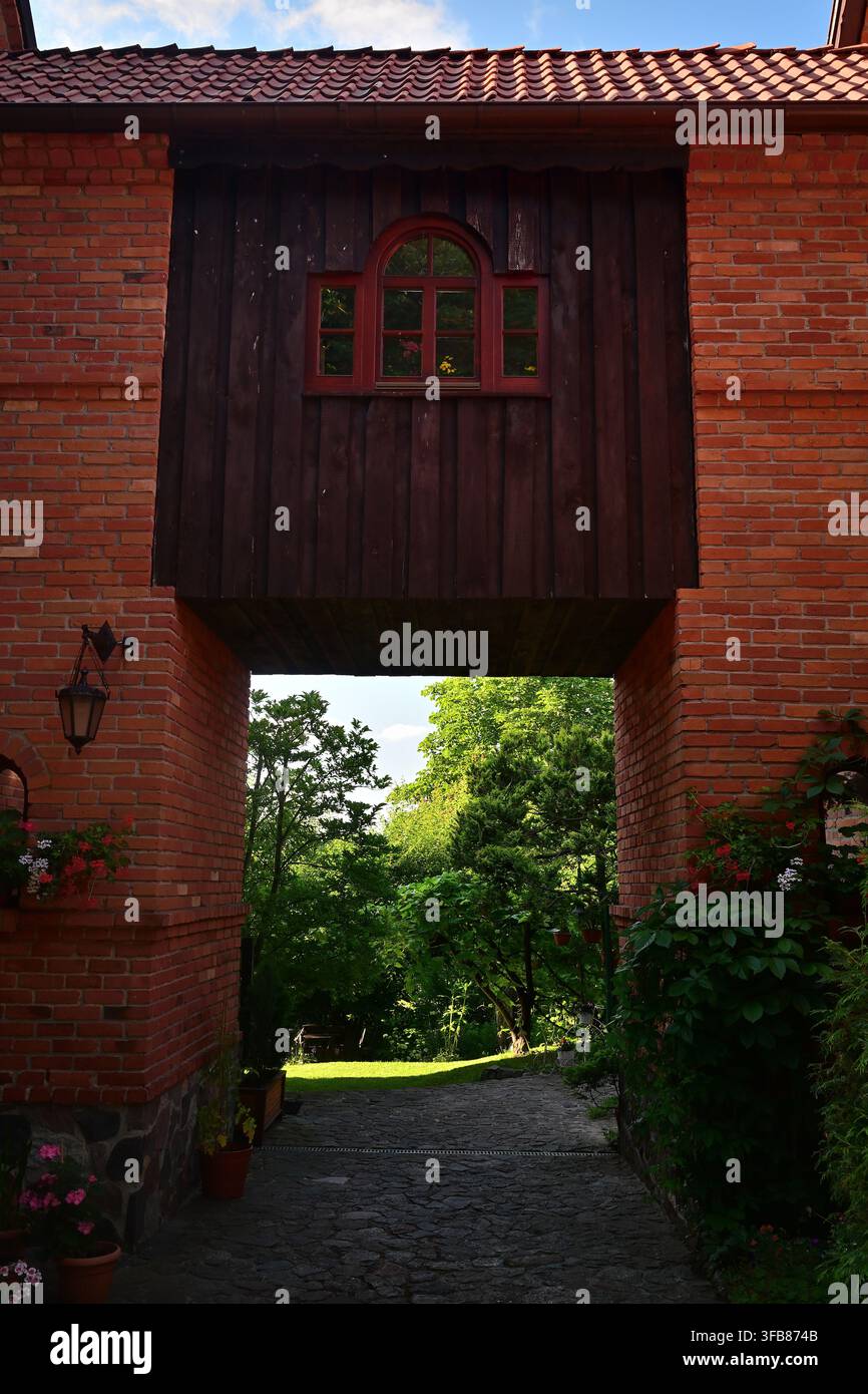 Une arche en brique avec une fenêtre en bois rouge sous laquelle passe un passage et une vue sur le chemin du jardin luxuriant. Banque D'Images