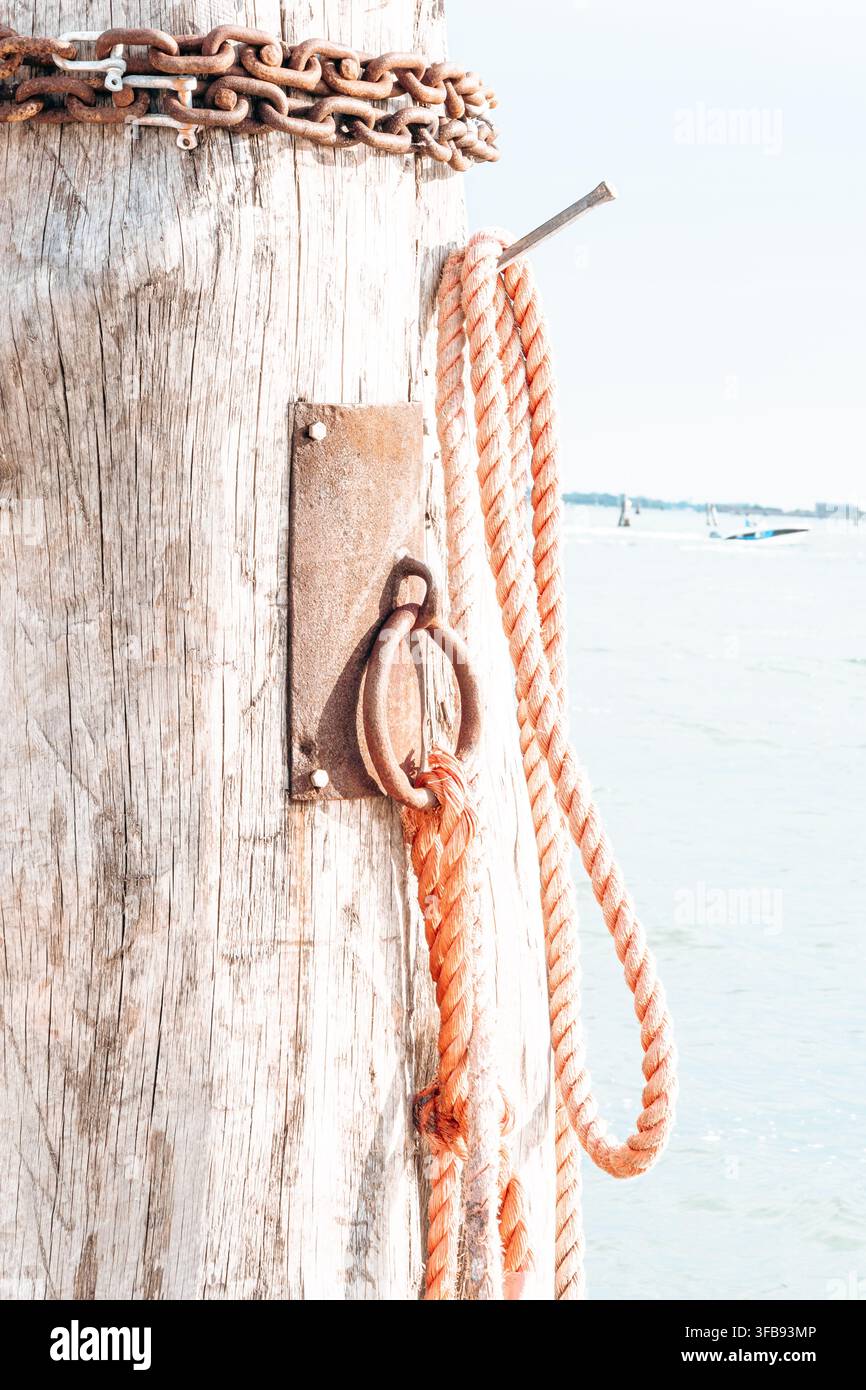 Une chaîne rouillée et des cordes d'amarrage orange pendent d'un ponton en bois patiné à côté d'une eau calme. Banque D'Images