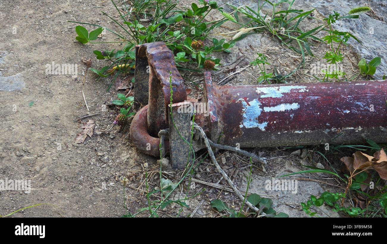Gros plan de composants industriels lourds en métal rouillé et d'une section de tuyau rouge écaillant abandonnée sur un sol sec avec des mauvaises herbes Banque D'Images