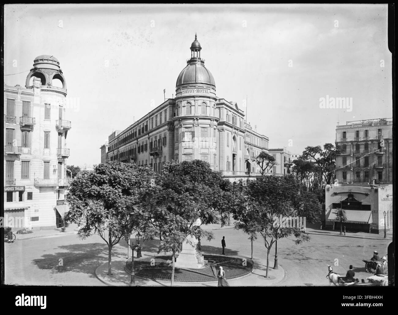 Photographie de 1910 du Savoy Hotel dans un cadre urbain au Caire, en Égypte, avec une enseigne indiquant « LADIES TAILOR / SAVOY-HOTEL », montrant le paysage urbain de l'époque, notamment la Place Soliman-Pasha. Banque D'Images