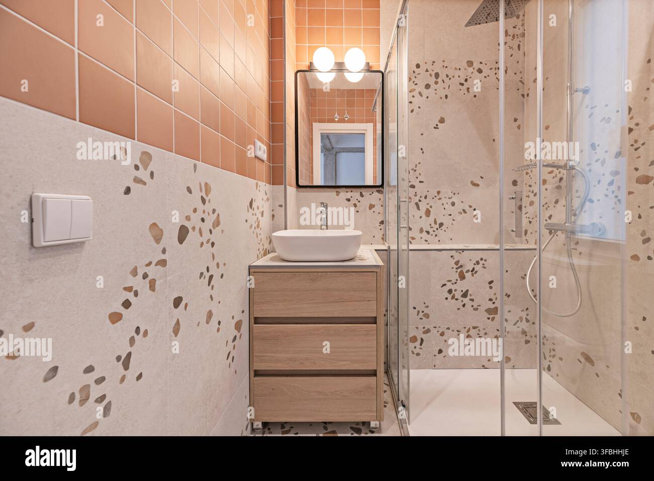 Salle de bain carrelée en terrazzo avec un mur d'accent en terre cuite, meuble de lavabo en bois, lavabo sur pied et cabine de douche à effet pluie vitrée. Banque D'Images