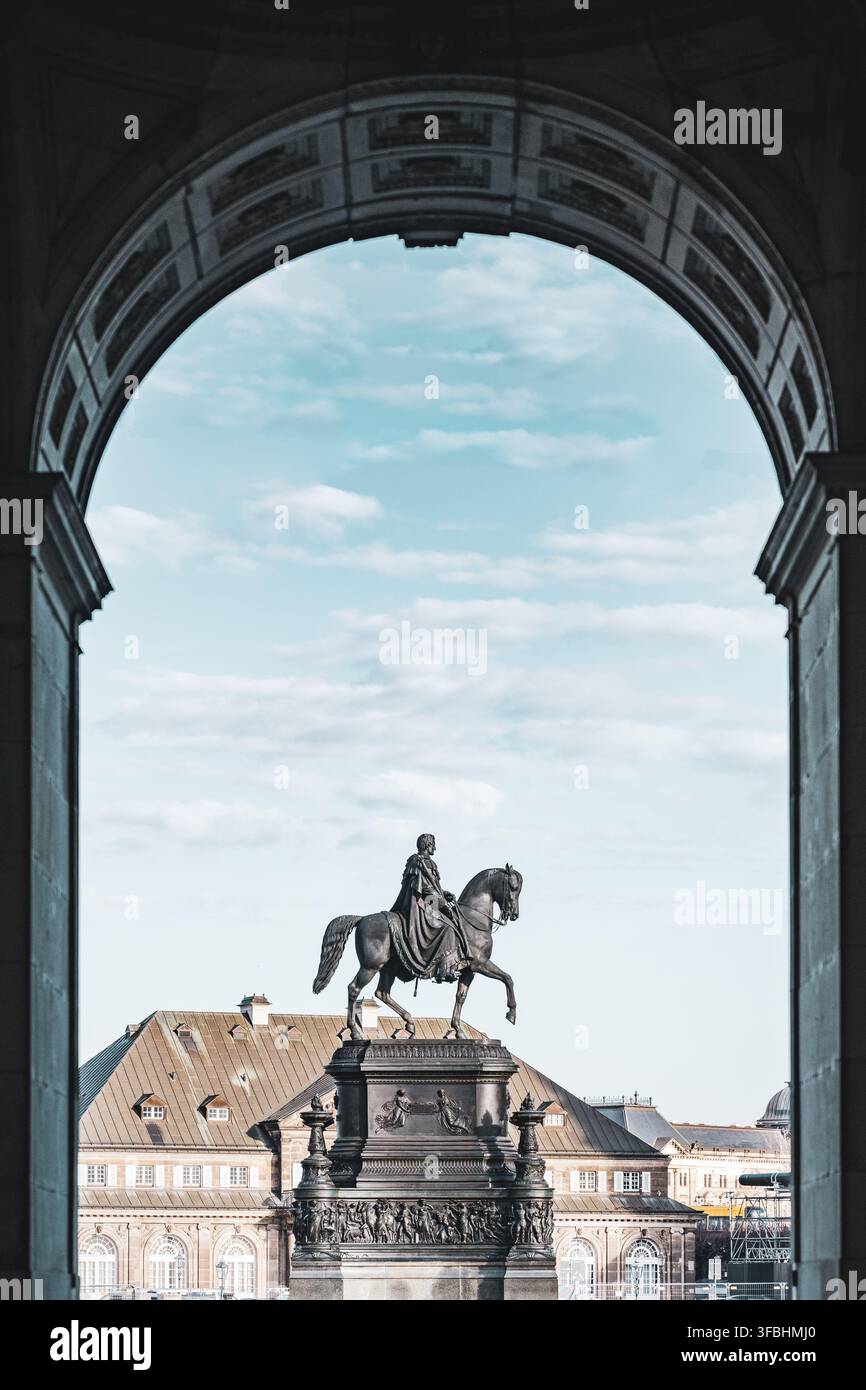 La statue du roi Jean se trouve à Dresde. Elle est placée sur un grand piédestal. Les gens peuvent la voir à travers une arche. Le ciel est clair et bleu au-dessus des bâtiments de la ville. Banque D'Images