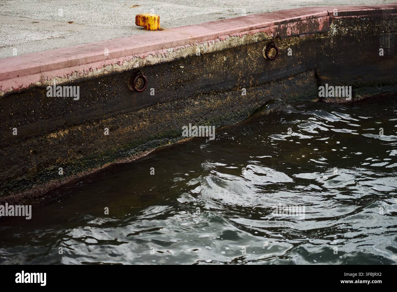 Vue rapprochée d'un quai en béton usé avec des anneaux métalliques rouillés et un petit taquet le long du bord de l'eau sombre en Grèce. Banque D'Images