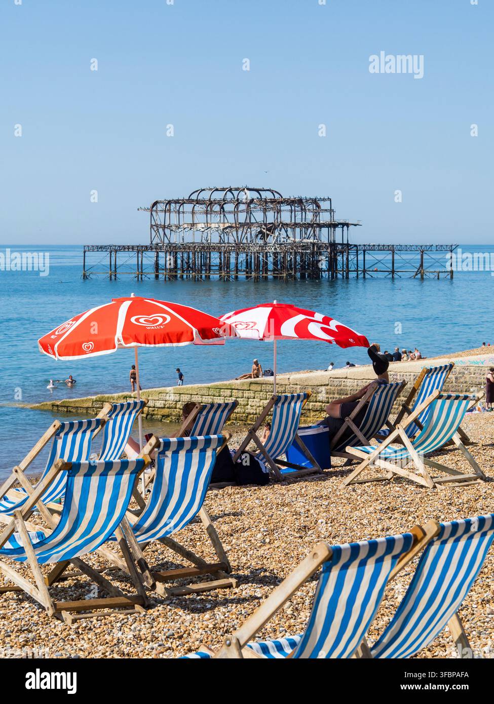 Les gens trouvent de l'ombre sous des parasols solaires, Canicule, avec le West Pier en ruine, Brighton Beach, Brighton, East Sussex, Angleterre, Royaume-Uni, GB. Banque D'Images