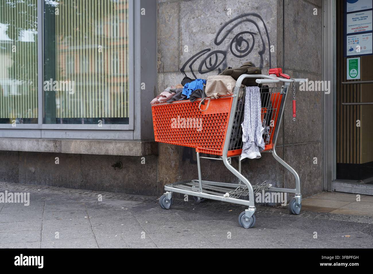 Berlin, Allemagne – 24 août 2026 : Un chariot de courses orange rempli de vêtements et d’objets personnels est abandonné à Hermannplatz à Neukölln. Banque D'Images