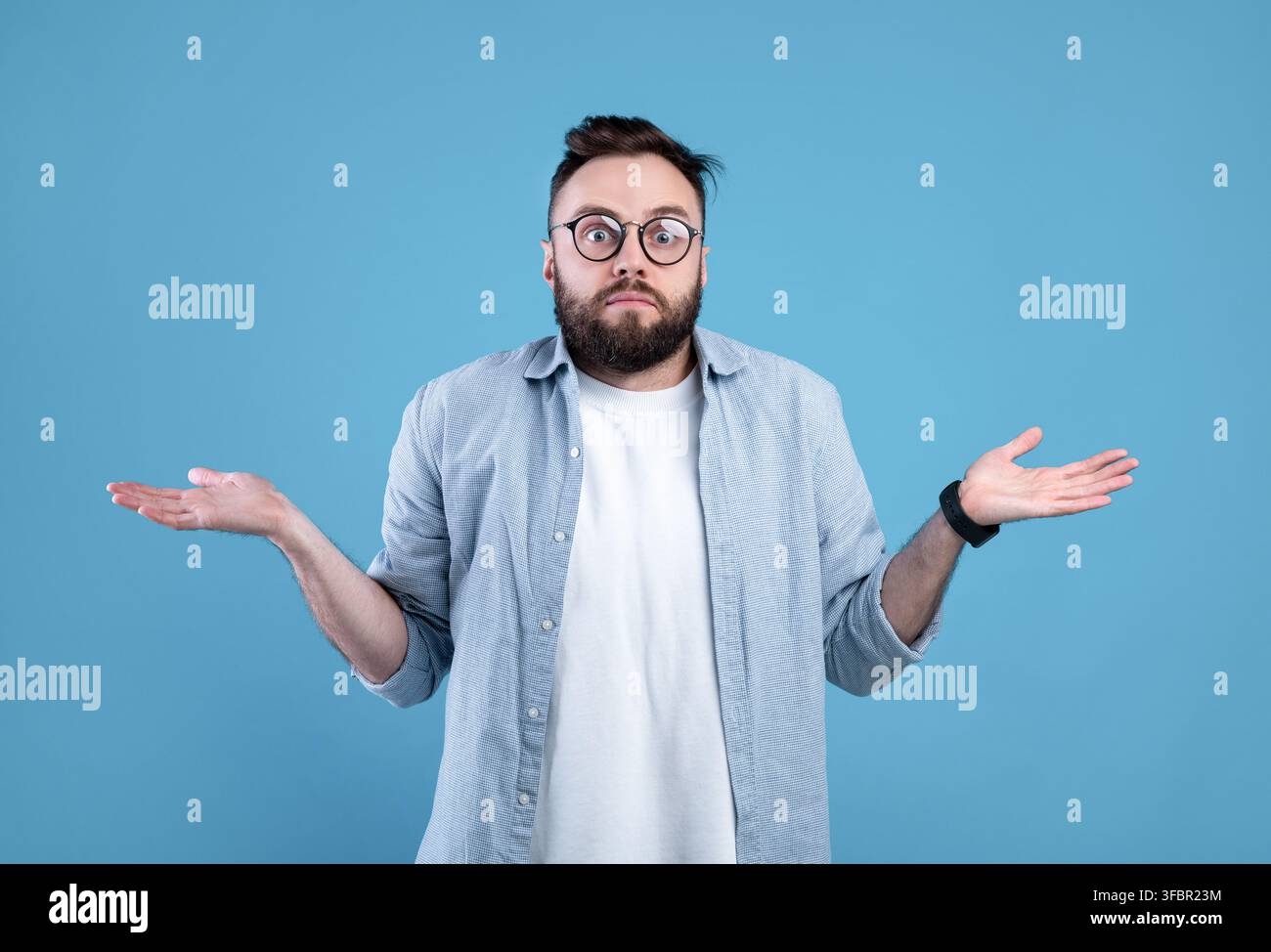 Qui sait. Homme barbu dans des lunettes haussant les épaules sur fond bleu studio Banque D'Images