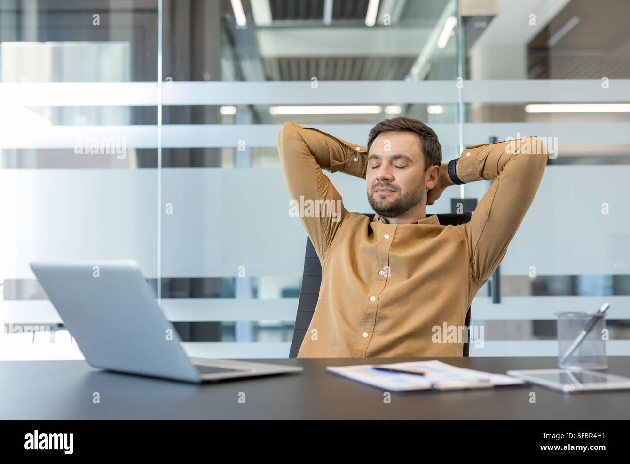 Homme d'affaires les yeux fermés se relaxant dans un fauteuil de bureau, prenant une pause paisible à son bureau avec ordinateur portable et documents, profitant d'un moment calme et attentif pendant la journée de travail Banque D'Images