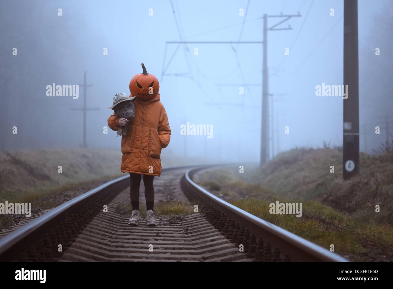 Personne avec une tête de citrouille sculptée debout sur des voies ferrées brumeuses tenant une tête de épouvantail. Portrait créatif d'Halloween avec une ambiance surréaliste et un paysage automnal Banque D'Images