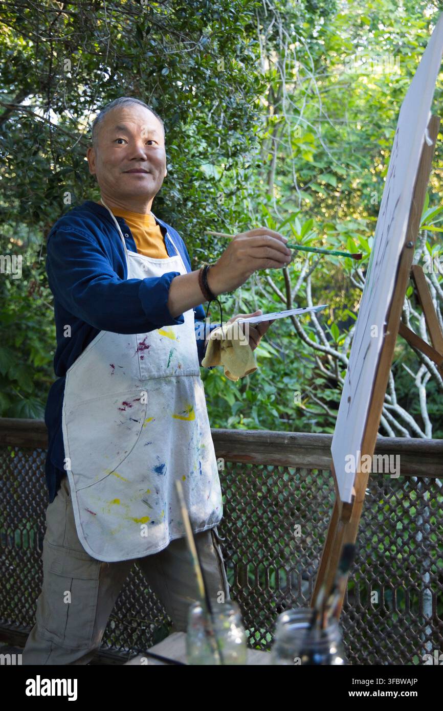 Homme senior asiatique portant un tablier taché de peinture, peignant sur le pont avec un chevalet, des pots de pinceaux et une palette Banque D'Images