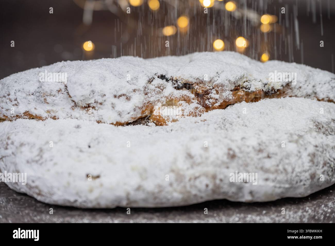 Un gros plan d'un stollen traditionnel aux fruits de Noël généreusement saupoudré de sucre en poudre tombant sur un fond sombre avec Banque D'Images