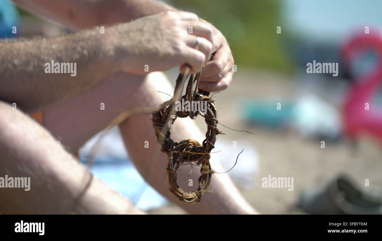 Close-up des mains d'un homme sur la plage créant une chaîne de cercles à partir de racines et de vignes tordues tout en se relaxant sur le sable pendant un été tranquille Banque D'Images