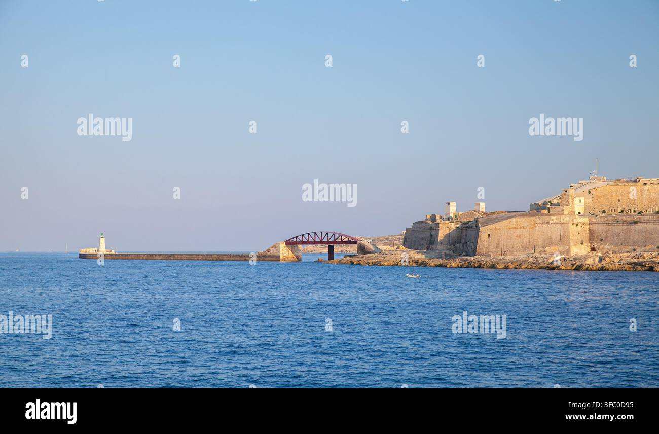 Une forteresse en pierre et un phare sont situés le long d'une longue jetée et d'un brise-lames tandis qu'un petit bateau dérive sur une eau bleue calme sous un ciel clair du soir. La Valette, Malte Banque D'Images
