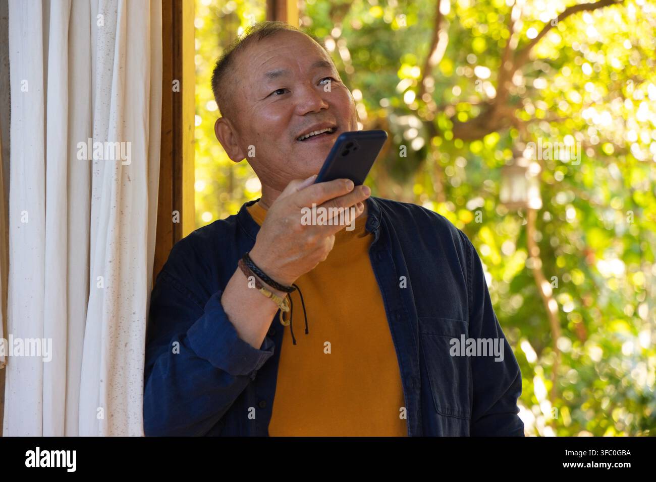 Homme asiatique âgé debout dans l'encadrement de la porte tenant un smartphone à la bouche, portant des bracelets, dans un jardin baigné de soleil Banque D'Images