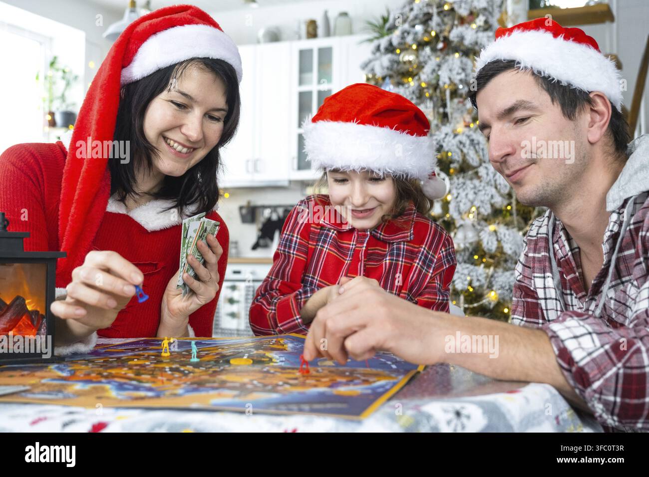 Famille traditionnelle heureuse composée de papa, maman et fille portant des chapeaux de Père Noël festifs, jouant à un jeu de société à table dans une cuisine blanche avec un sapin de Noël et des décorations. N Banque D'Images