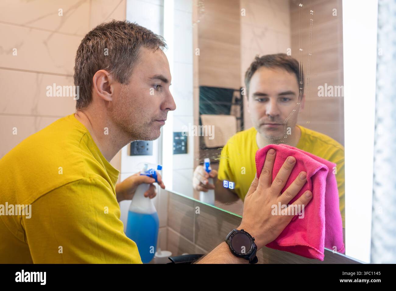 Homme nettoyant un miroir de salle de bain avec un chiffon rose et une bouteille de pulvérisation, regardant son reflet. Ménage, hygiène et routine de nettoyage à la maison. Banque D'Images
