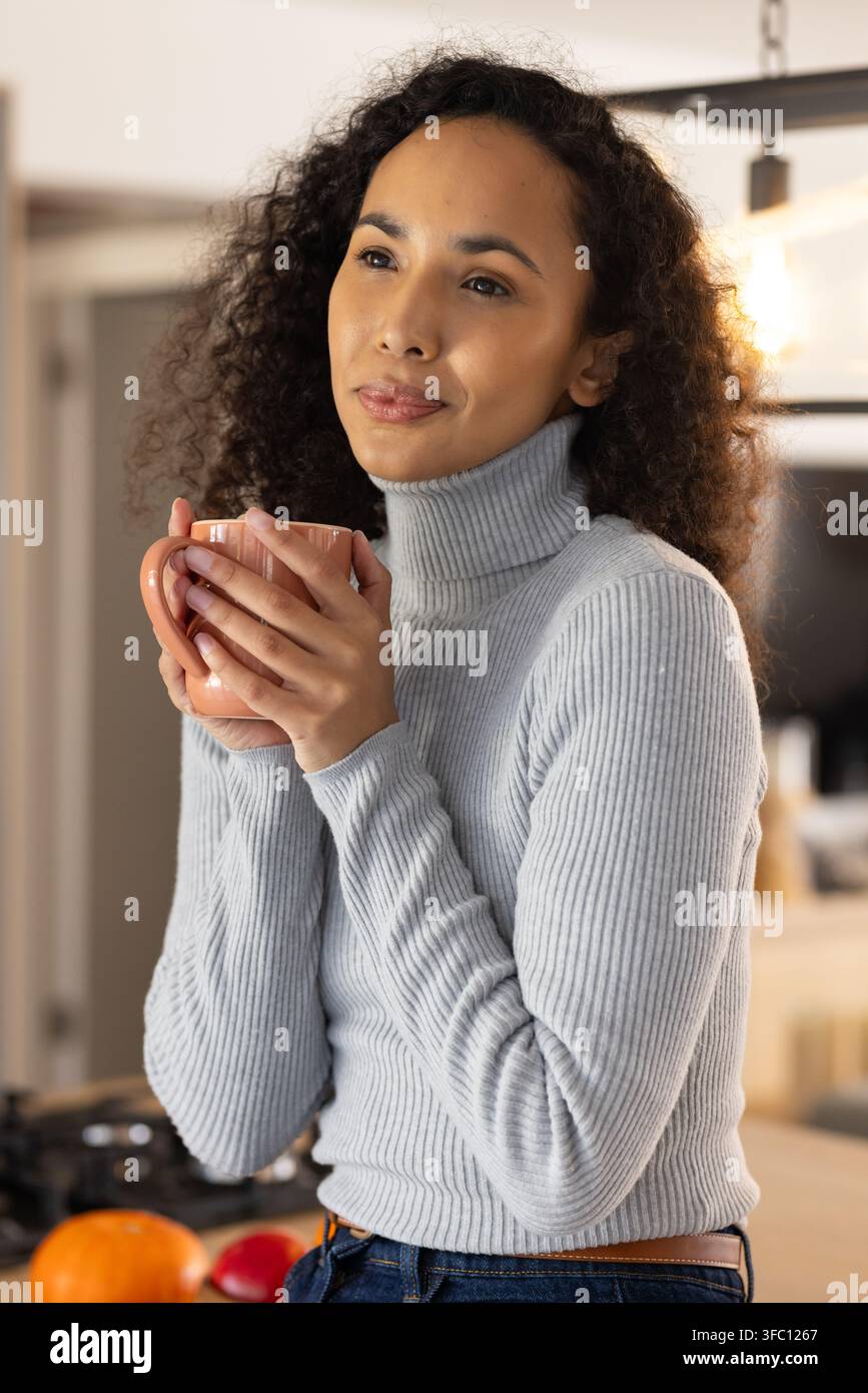 Femme afro-américaine portant un pull gris clair tenant une tasse et regardant dans la cuisine de la maison Banque D'Images
