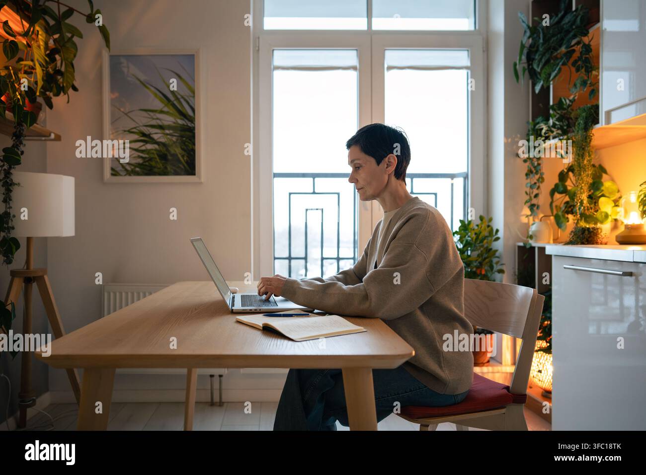 Femme mûre freelance travaillant sur un ordinateur portable dans un intérieur confortable avec des plantes d'intérieur et un éclairage chaleureux Banque D'Images
