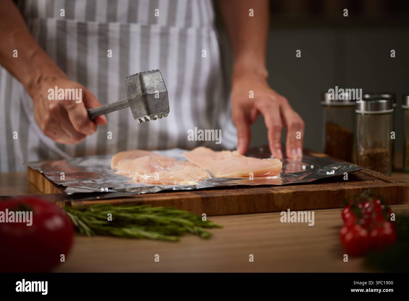Cuisinier portant un tablier préparant des filets de poulet crus sur du papier d'aluminium avec un marteau à viande dans un cadre de cuisine lumineux Banque D'Images