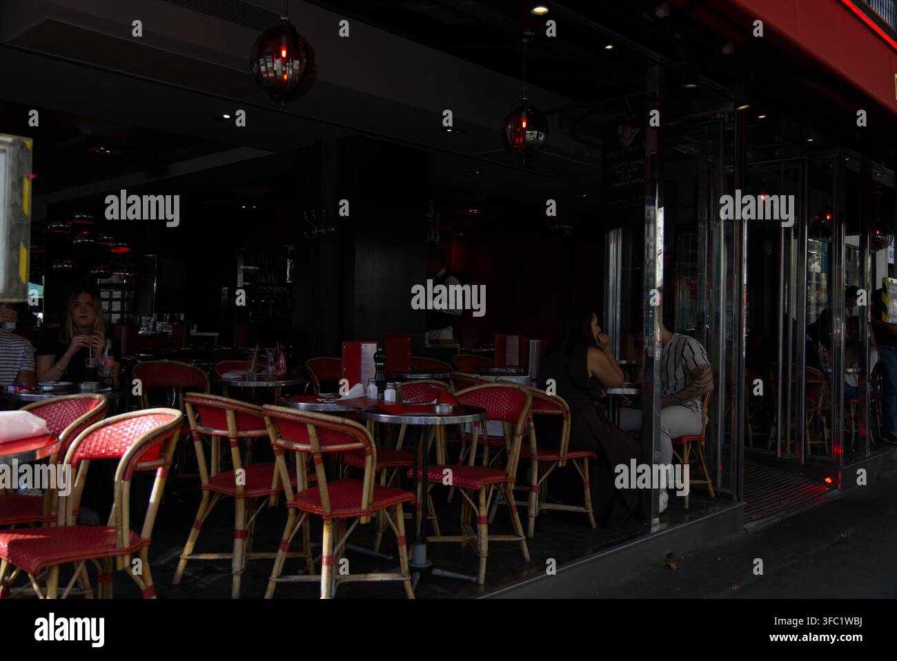 Clients assis sur des chaises bistro rouges sur une terrasse de café en plein air avec un intérieur de restaurant sombre et atmosphérique en arrière-plan. Banque D'Images