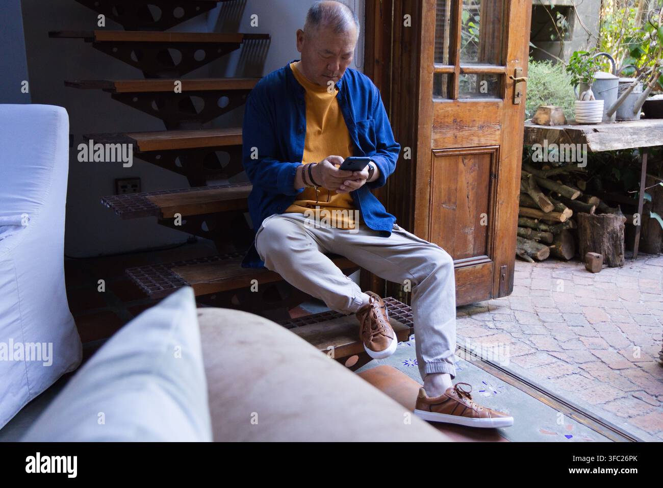 Homme asiatique âgé assis sur des marches surélevées à la maison, utilisant un smartphone près d'une porte en bois ouverte Banque D'Images