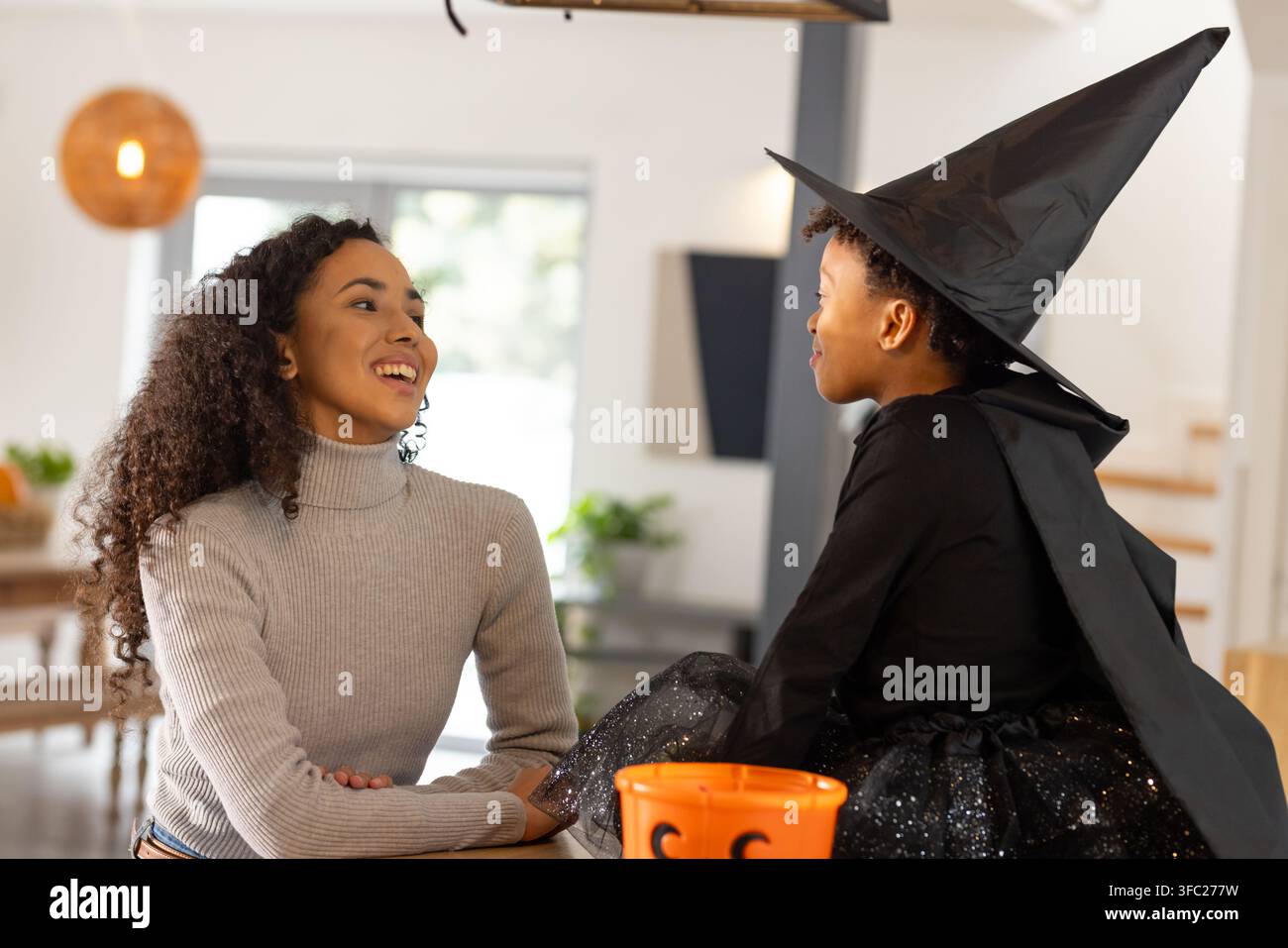 Mère et fille afro-américaines assises au comptoir, enfant en costume de sorcière tenant un seau à citrouilles Banque D'Images