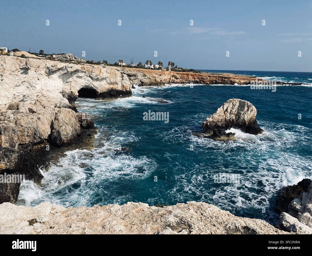 Des vagues blanches se déplacent à travers une eau bleu profond sous des falaises côtières pâles, avec une petite grotte marine et des rochers érodés le long du rivage. - Image de stock capturée avec un smartphone