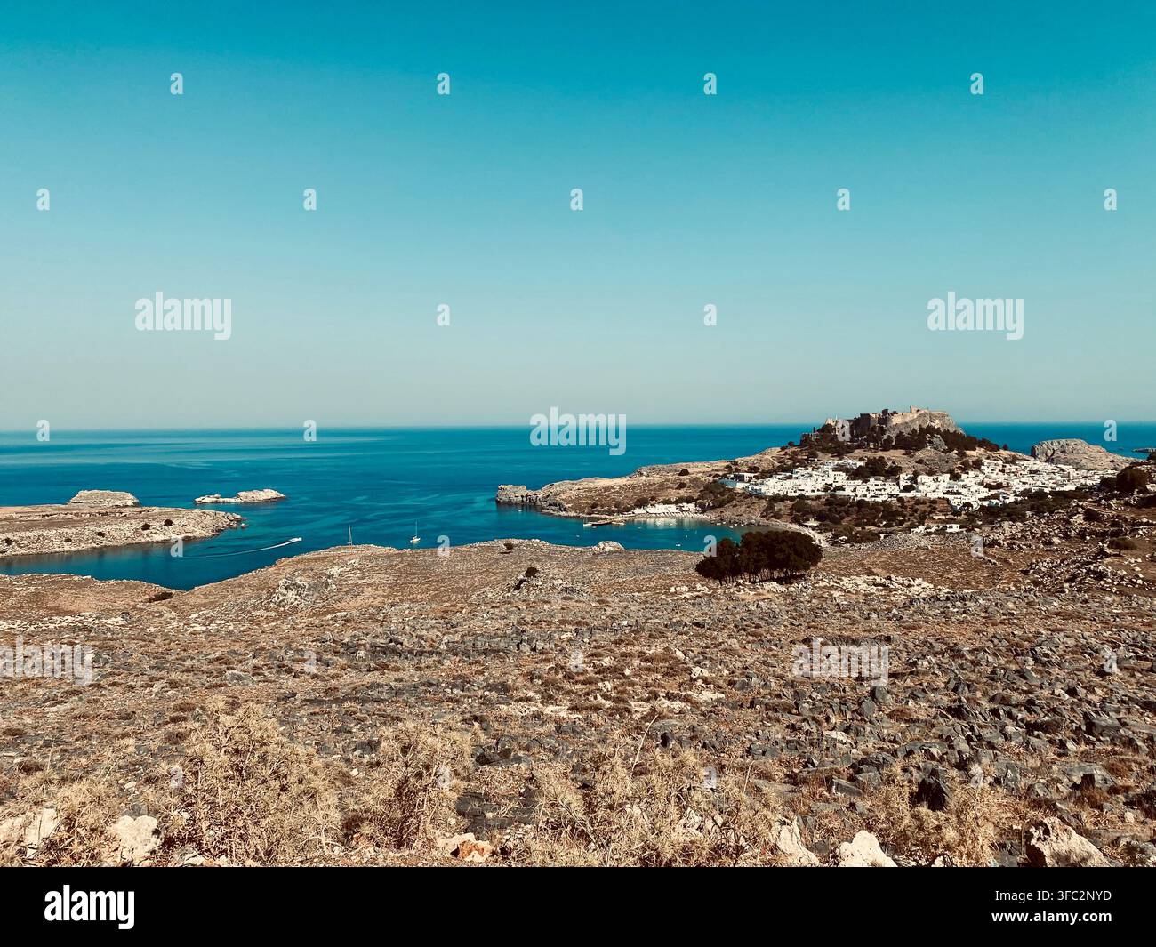 Un village côtier blanchâtre se trouve en contrebas de ruines au sommet d'une colline près d'une baie bleue et calme, entouré d'un terrain rocheux aride. - Image de stock capturée avec un smartphone