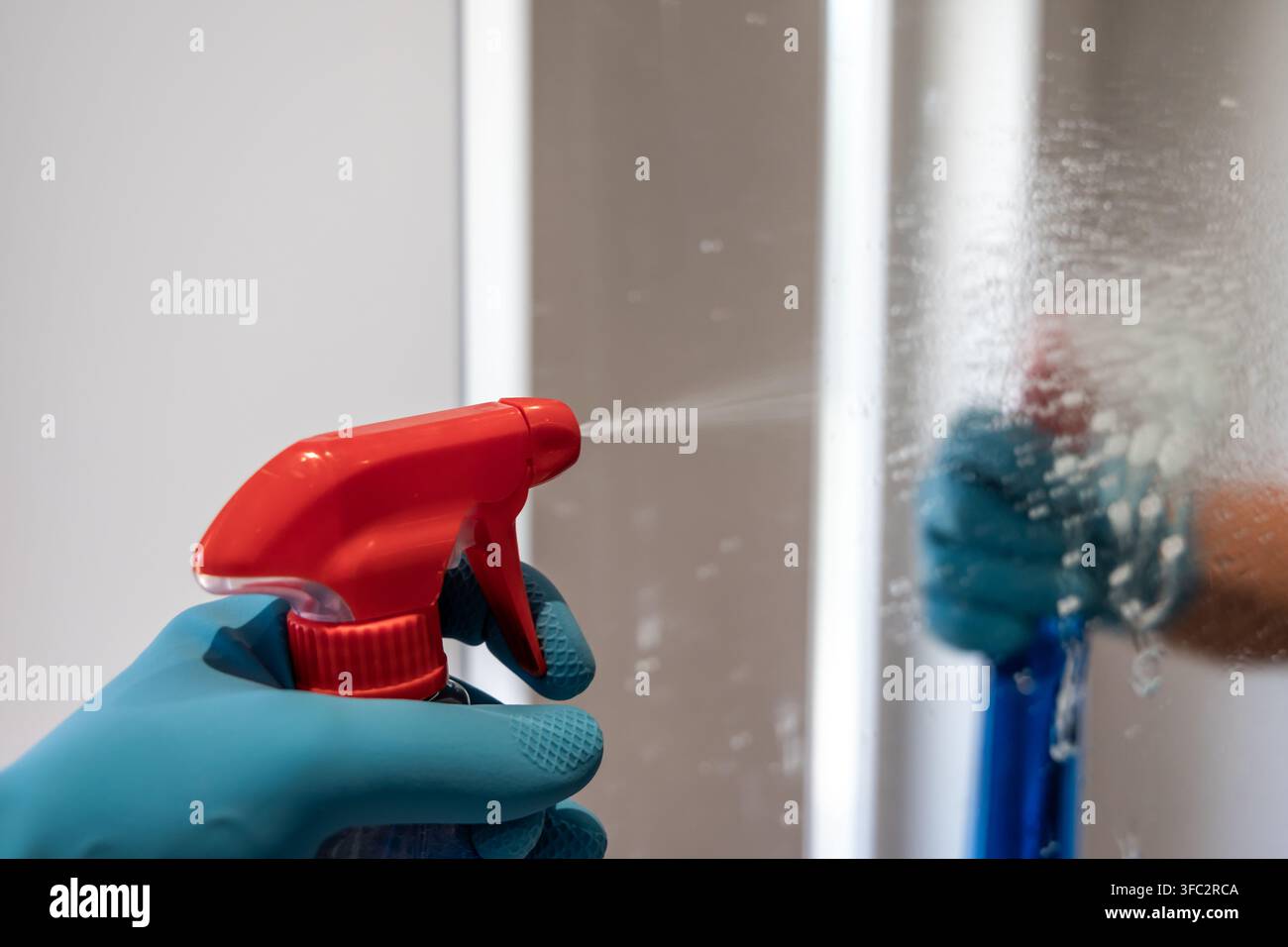 Gros plan d'une main portant un gant bleu tenant une bouteille de pulvérisation rouge et vaporisant une surface vitrée avec une solution de nettoyage Banque D'Images