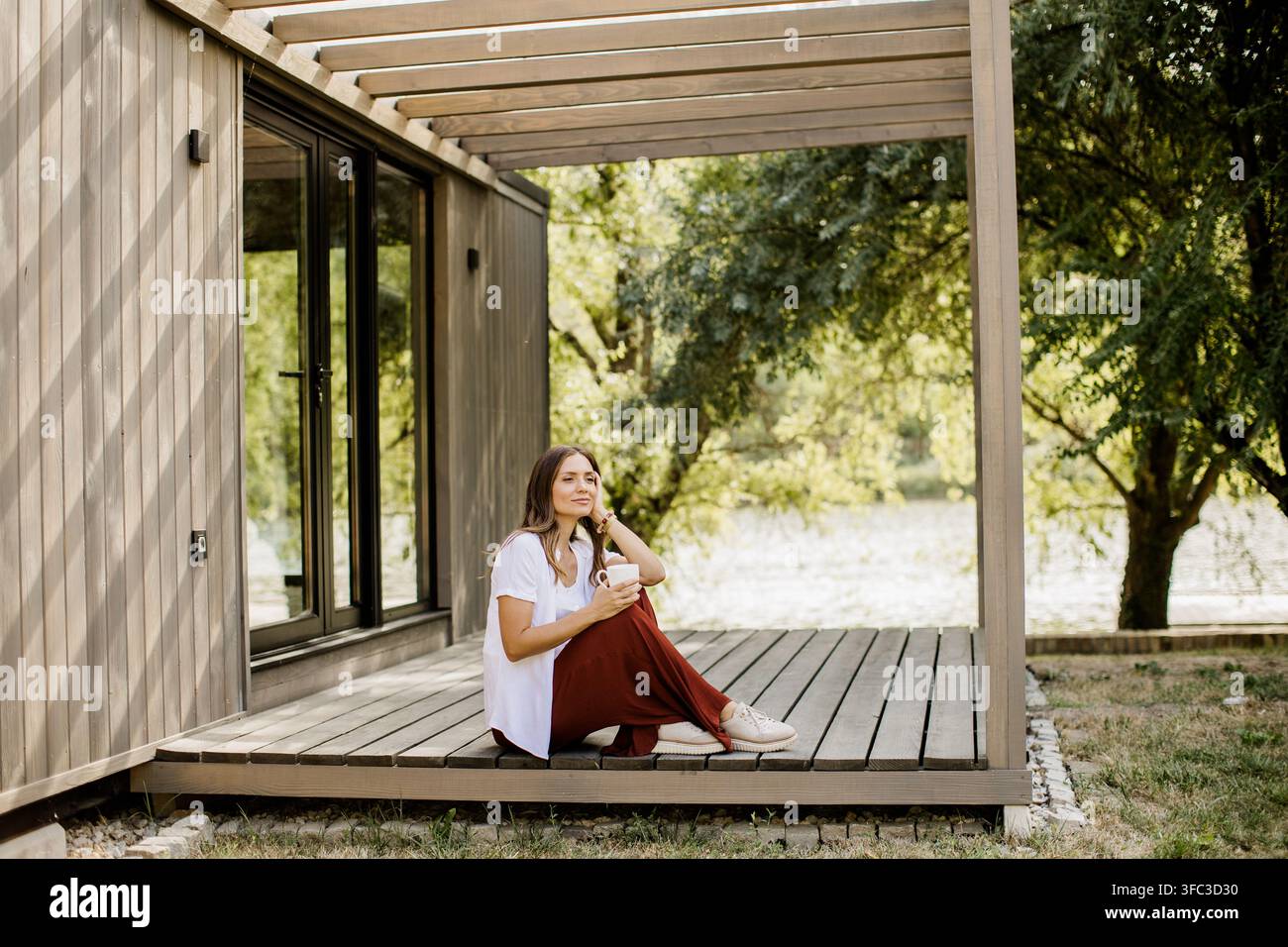 Une jeune femme se détend sur une terrasse en bois, sirote son café tout en profitant de la vue sereine sur la rivière entourée de verdure. Banque D'Images